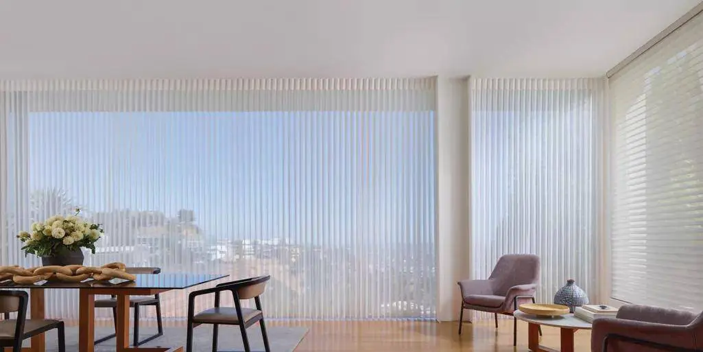 Spacious living space with large windows featuring vertical blinds, minimalist furniture, cozy armchair, and decorative accents, perfect for stylish, natural light-filled interiors.