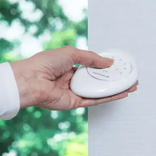 A hand holding a white security sensor device against a blurred green outdoor background.