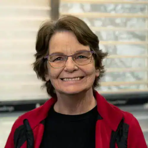 Sandi Switzer smiling woman with glasses, wearing a red jacket, indoors with window blinds in background.