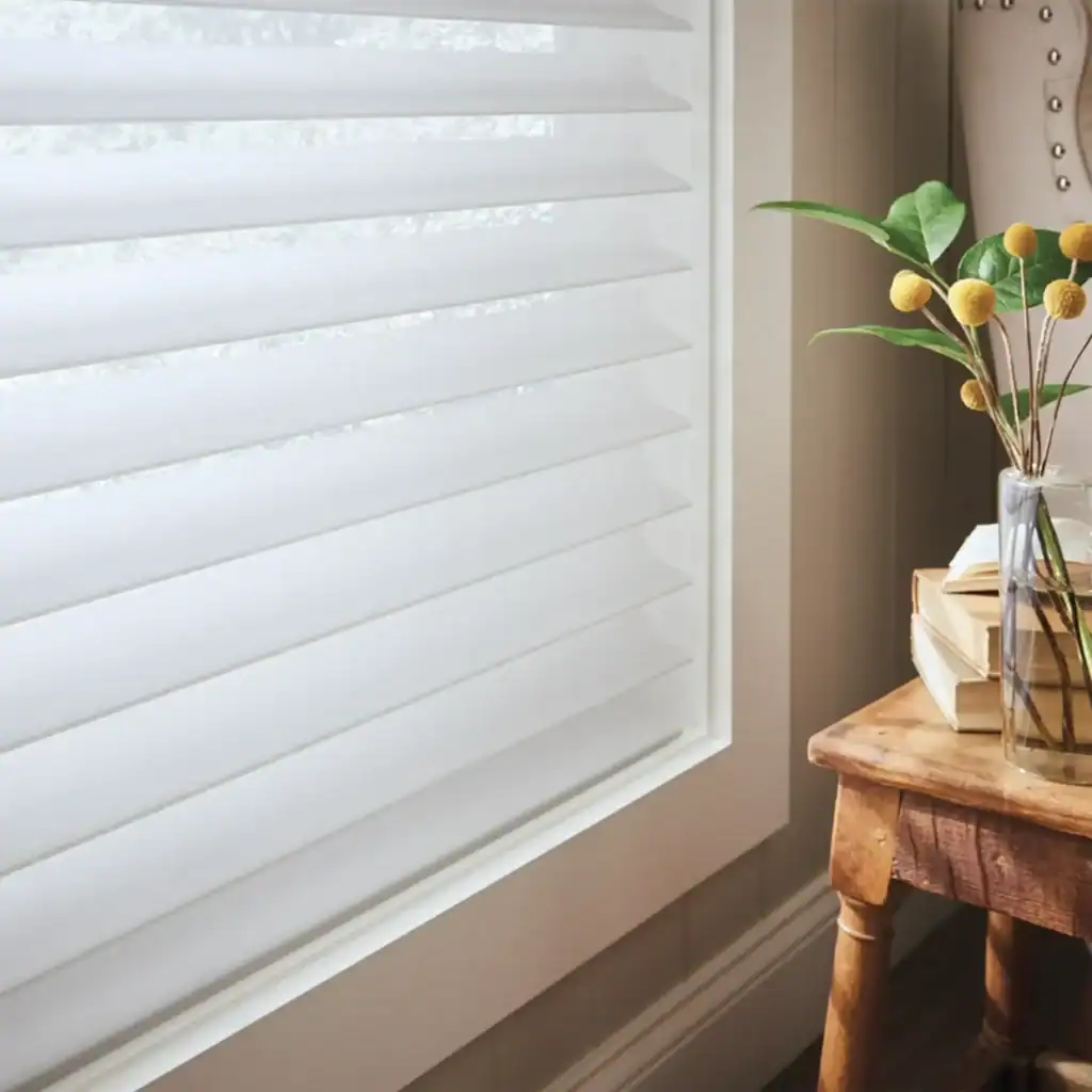 Bright window with white horizontal blinds, natural light, and a glass vase with green leaves and yellow flowers on a rustic wooden table.